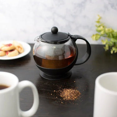The Steep in Style Tempered Glass Teapot with Stainless Steel Infuser sits on a dark table beside two white mugs, scattered loose-leaf tea, a blurred plate of pastries in the background, and a plant on the right.